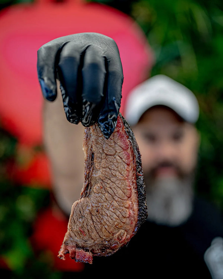 A thick slice of cooked beef held up on a gloved hand, dark bark down one edge, the cook out of focus behind
